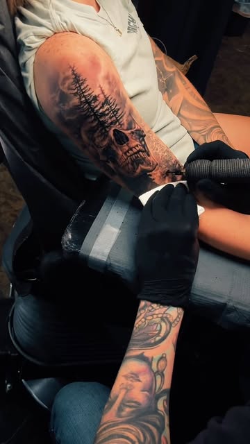 Tattoo session showing a skull and pine forest composition in progress on an upper arm