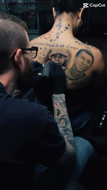Tattoo artist working on a large black-and-grey memorial portrait composition across a client’s back